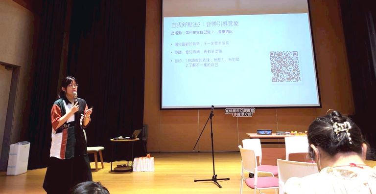 Amanda Lee giving a presentation in a dimly lit conference room, with a screen displaying text and a QR code. Audience members are seated in the foreground.