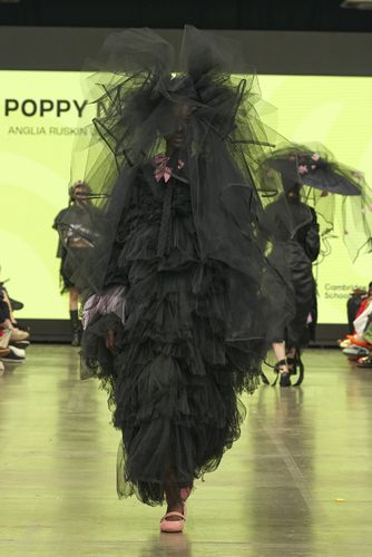 Model in a voluminous black ruffled dress and large hat walks on a runway, with a bright green screen in the background.