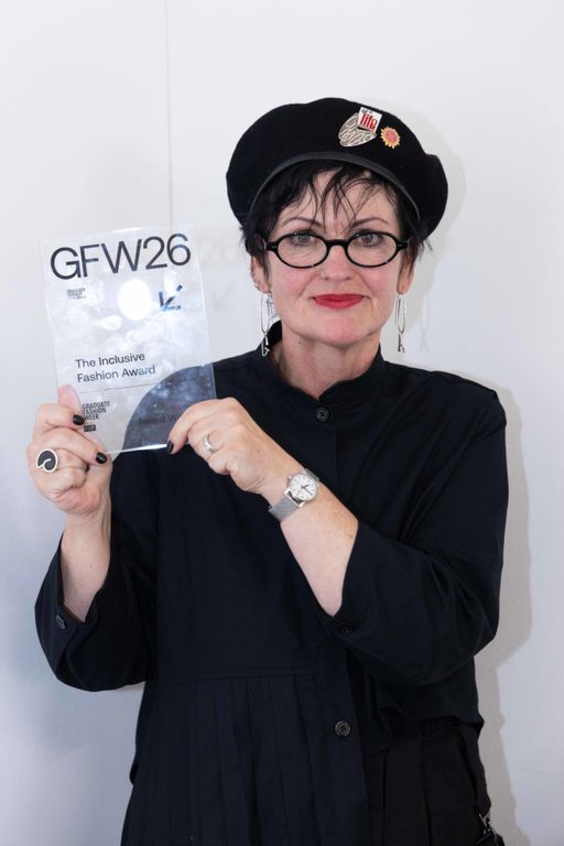 Gunta Andrews in a black beret, glasses and top holding up her Inclusive Fashion award from Graduate Fashion Week