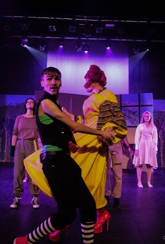 Actors in colorful costumes dance energetically on a stage with dramatic lighting and a backdrop of trees.