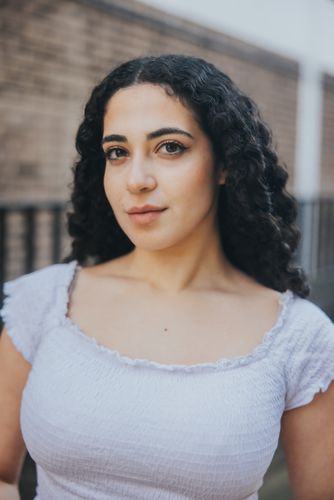 A woman with curly hair and a white top looks directly at the camera against a blurred brick background.