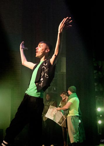 Male actor on stage with arms raised, wearing a vest and green t-shirt; actors in the background hold a newspaper, smiling.