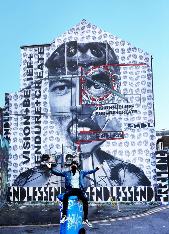 A large mural of a person cut-up on the side of a house, with Endless sitting on a recycling bin below