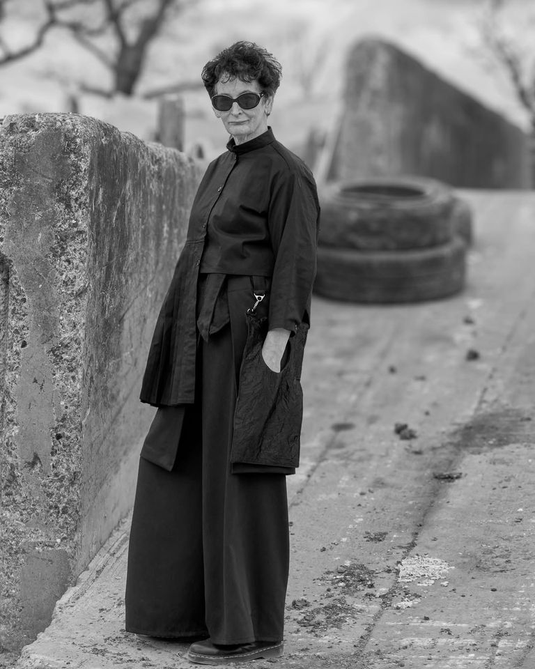 Black and white photo of female model wearing black sunglasses and trouser suit with industrial background