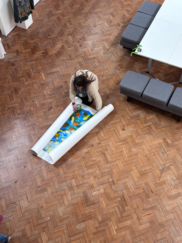 A person kneels on a wooden floor, taping a colorful rolled-out poster, with tables and gray cushions nearby.