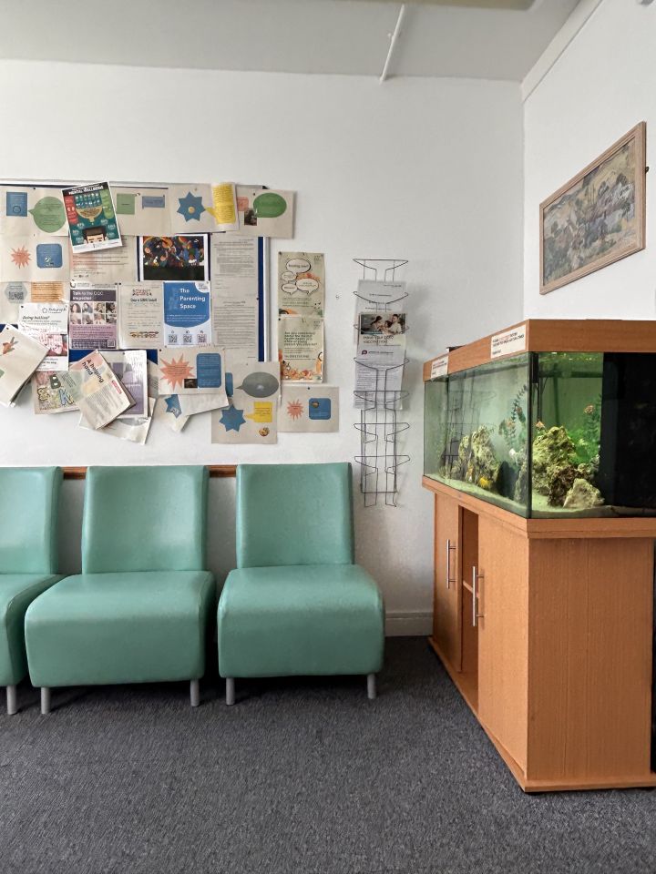 A waiting room with teal chairs, a bulletin board filled with papers, a wall-mounted magazine rack, and a fish tank on a wooden stand.