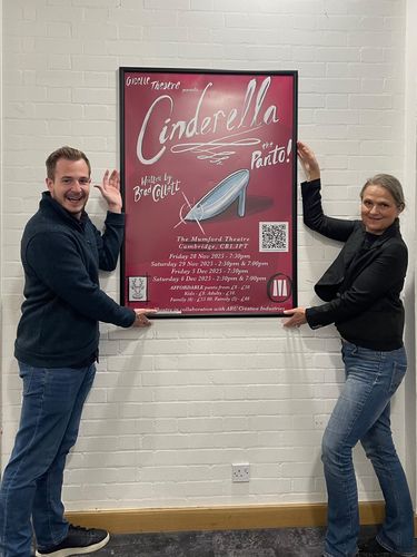 Brad Collett and Susan Hedgecoe hold a "Cinderella" poster on a white brick wall. The poster features a glass slipper and show details.