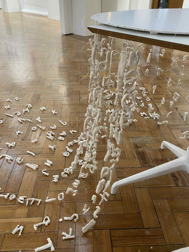 Sculpture made from strings of white letters suspended from disc, draped over table