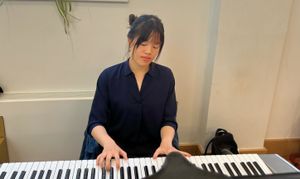 Amanda Lee playing a keyboard with closed eyes, seated in a room with a white wall and a hanging plant.