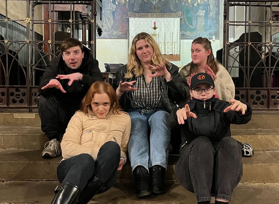 Five people sitting on stairs, playfully posing with claw-like hand gestures in front of an ornate wall background.