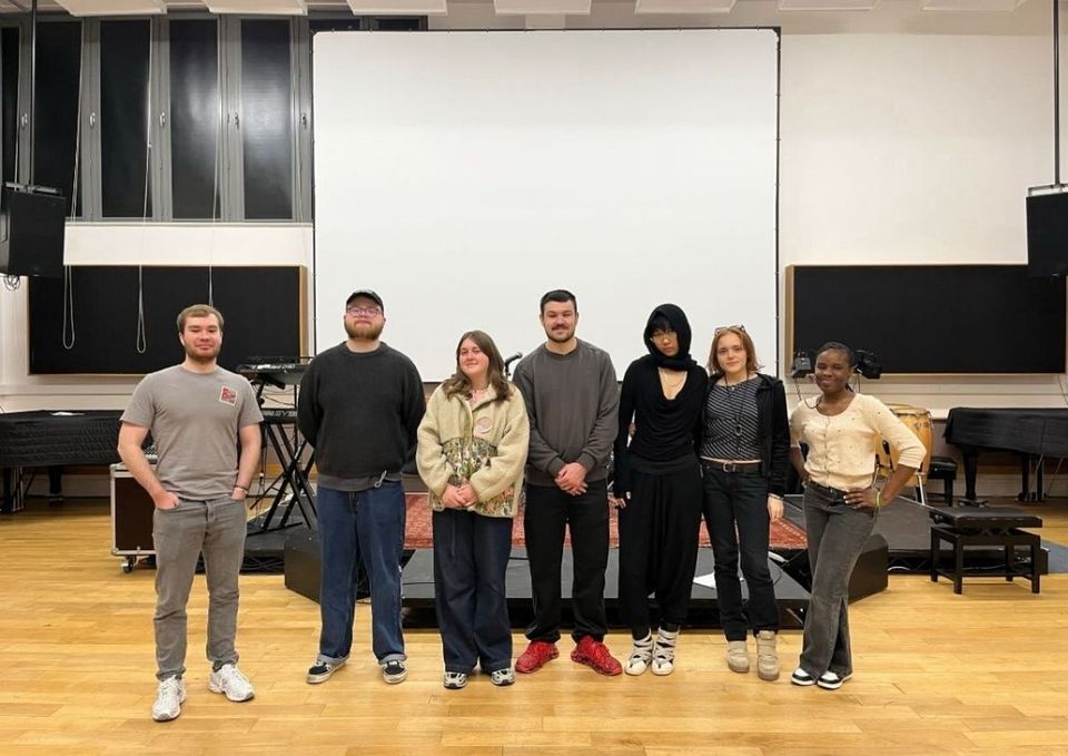 Seven people stand together in a music studio with instruments in the background, posing for a group photo.