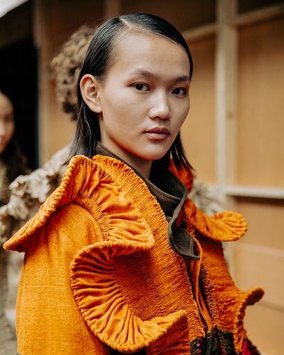 Model wearing a vibrant orange textured garment with unique ruffled details, standing indoors with a neutral expression.