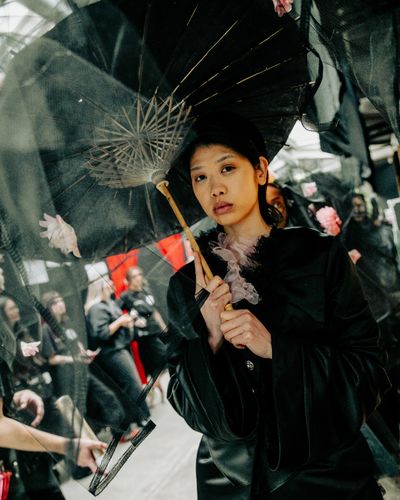 Woman in black holding a parasol adorned with flowers, standing in a crowd, wearing a thoughtful expression.