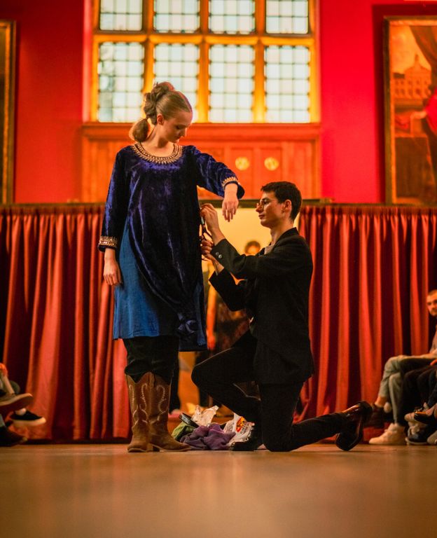Csomin Diaconu kneeling and adjusting a female model's dress on stage. She's in a blue dress and boots. The backdrop is red with a large window above.