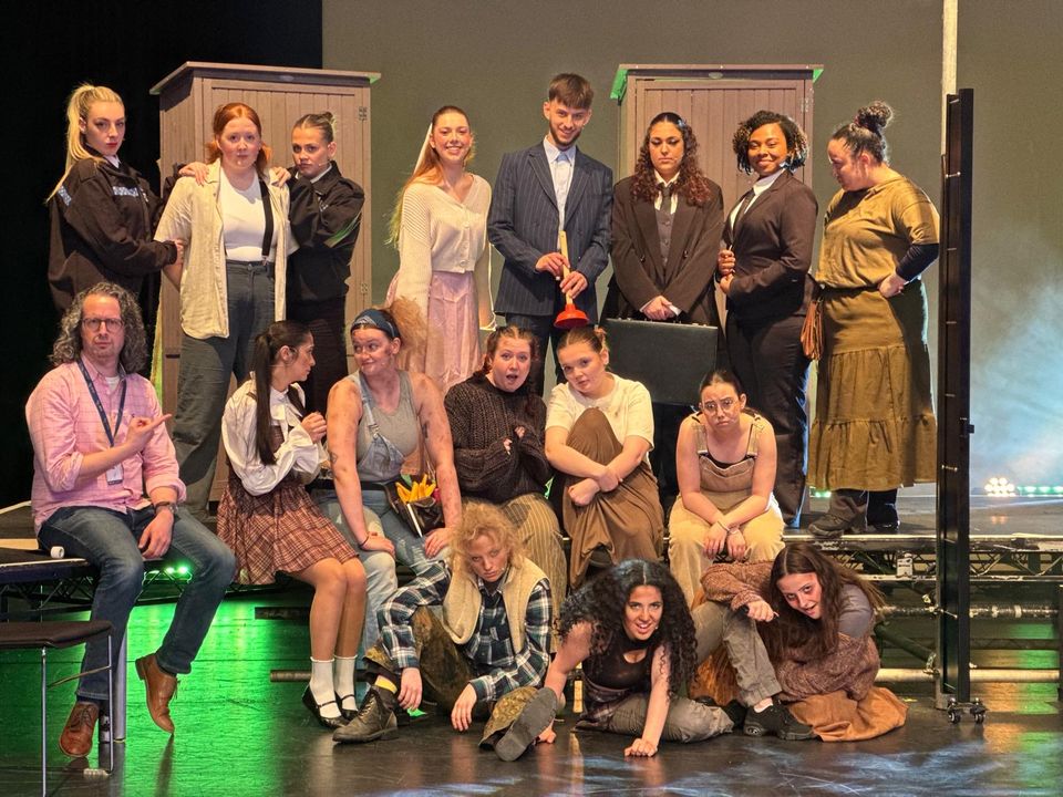 A diverse theatre cast poses on stage in character, showing various costumes and expressions, with a director seated to the side.