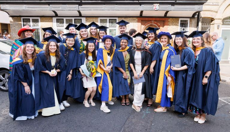 A group of graduates in blue robes and caps stand together, smiling and holding diplomas outside a building on a sunny day.