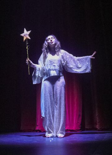 A female actor in a sparkling silver costume holds a star-topped wand, standing on stage with dramatic lighting and a red curtain backdrop.