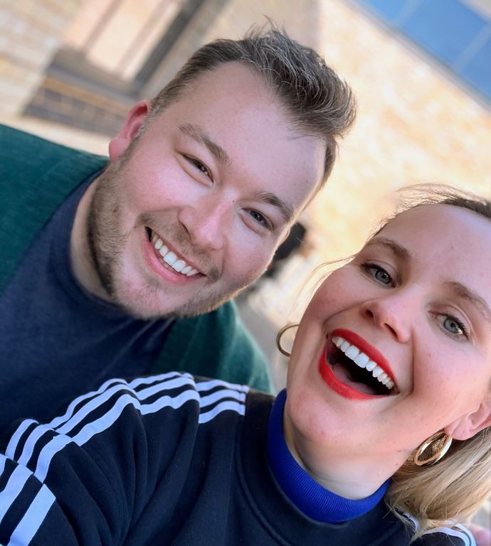 A smiling man and woman taking a selfie outdoors. The woman wears red lipstick and hoop earrings, and both look cheerful.