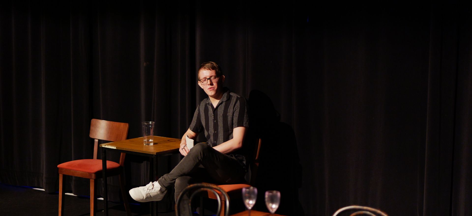 Jack Palmer sitting on stage on wooden chair, leaning on wooden table