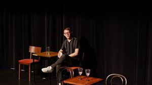 Jack Palmer sitting on stage on wooden chair, leaning on wooden table