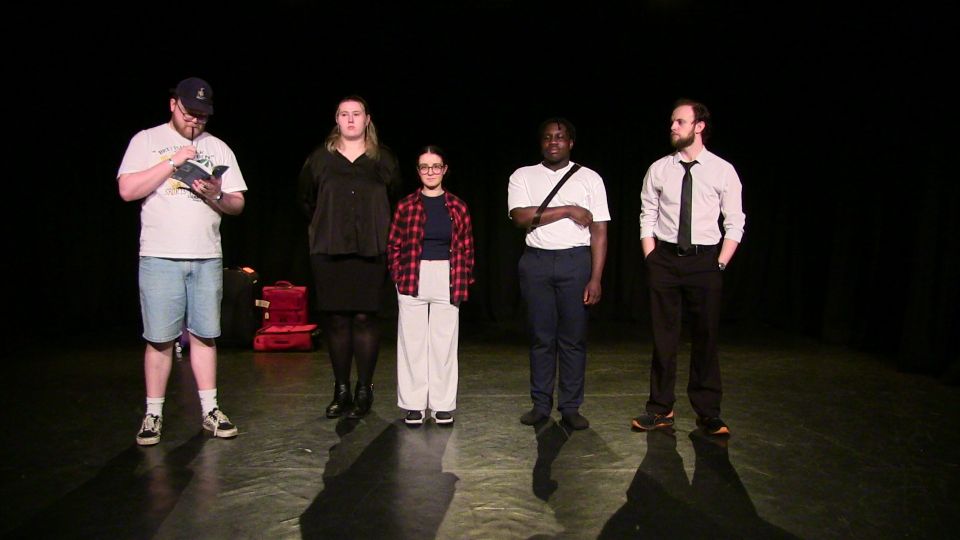 Five people stand on a dark stage with red suitcases in the background, each dressed in different casual and formal attire.