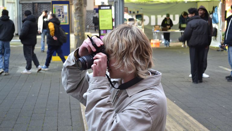 Person with blonde hair in a beige jacket taking a photo with a camera on a bustling street with people and stalls in the background.