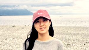 Charlee wearing a pink cap and gray sweater stands on a rocky beach, with mountains and a cloudy sky in the background.