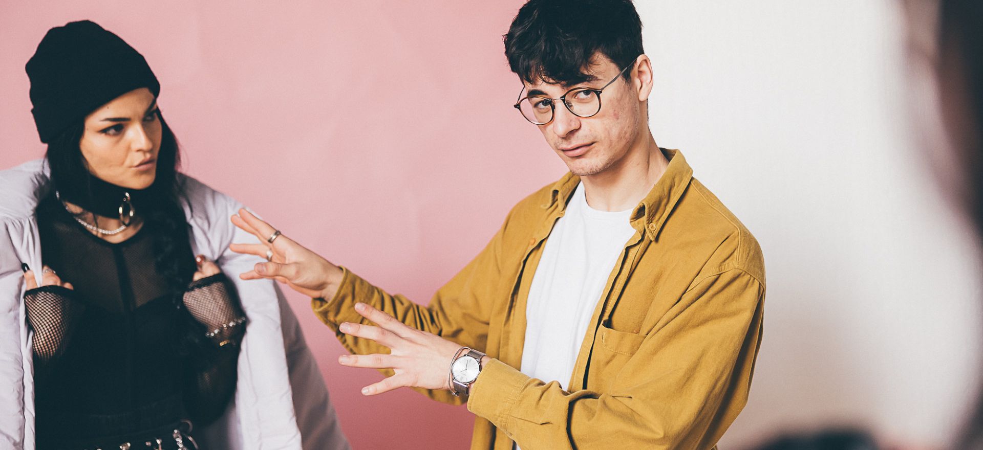 Cosmin Diaconu gestures while talking to a woman in a black beanie and edgy outfit against a pink and white background.