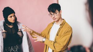 Cosmin Diaconu gestures while talking to a woman in a black beanie and edgy outfit against a pink and white background.