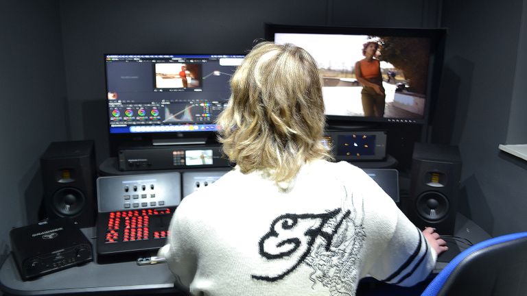 Jacob in a white sweater editing video on dual monitors, surrounded by audio equipment in a dimly lit room.