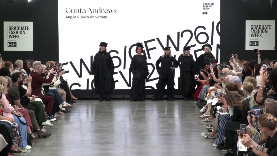 Five female models lined up in front of screen on GFW2026 catwalk wearing black costumes with black hats