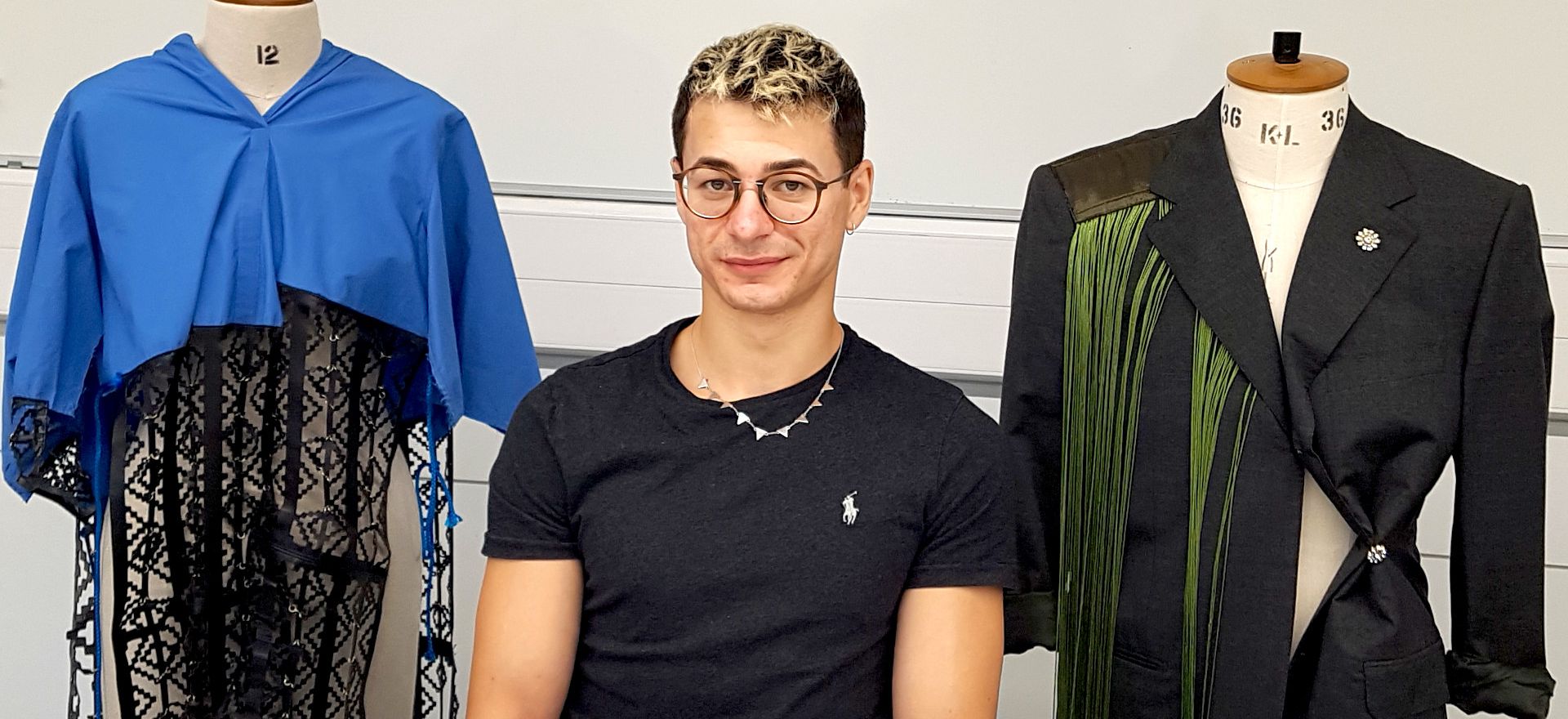 Cosmin Diaconu sitting between two mannequins displaying a blue garment with lace and a black coat with green accents.