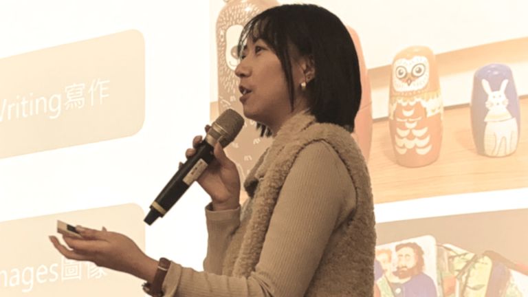 Charlee Tsai speaking into a microphone, gesturing with her hand, in front of a screen displaying text and images of nesting dolls.