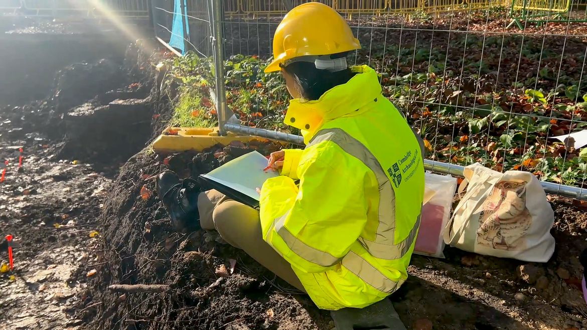 Photo of student in high-vis jacket and helmet drawing at dig site