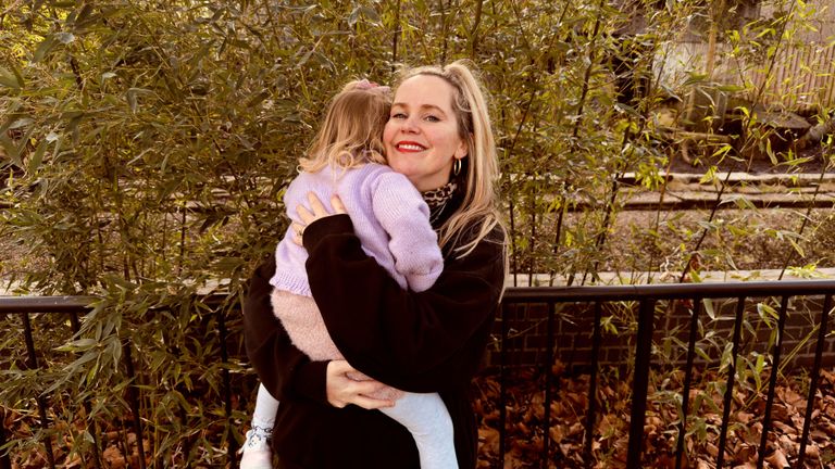 Emily MacRae hugs her daughter in a purple sweater, standing in front of leafy bushes and a rail fence.