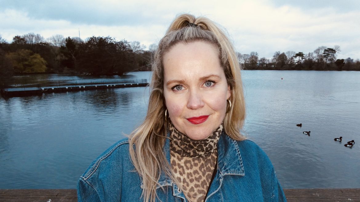 Emily McCrae wearing a denim jacket and leopard print scarf, stands by a lake with ducks swimming behind her.