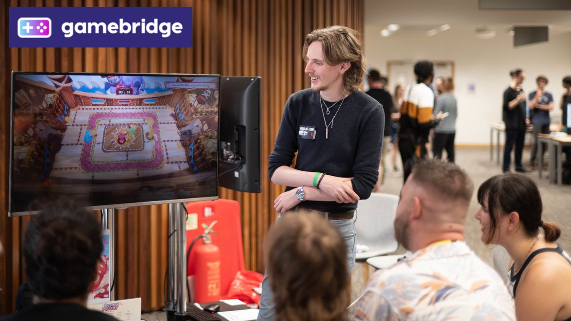 A group watches a video game on a screen. A person stands nearby, smiling and talking. A "gamebridge" logo is displayed in the corner.