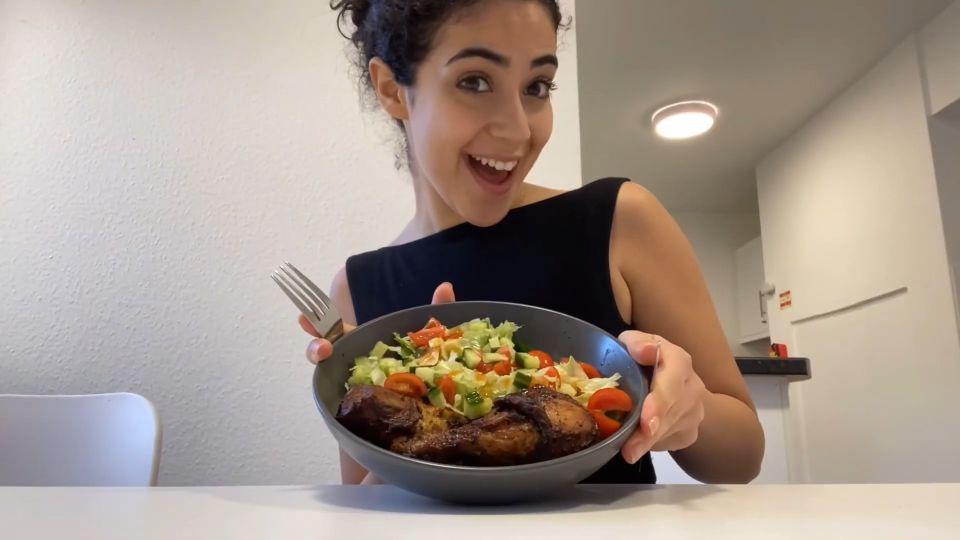 Smiling person holding a plate of grilled chicken and fresh salad, seated at a white table in a brightly lit room.