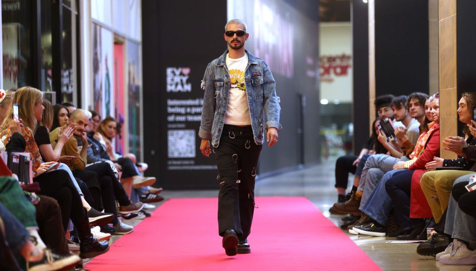 A male model with sunglasses walks confidently on a red carpet runway, wearing a denim jacket and black pants, with an audience seated on both sides.