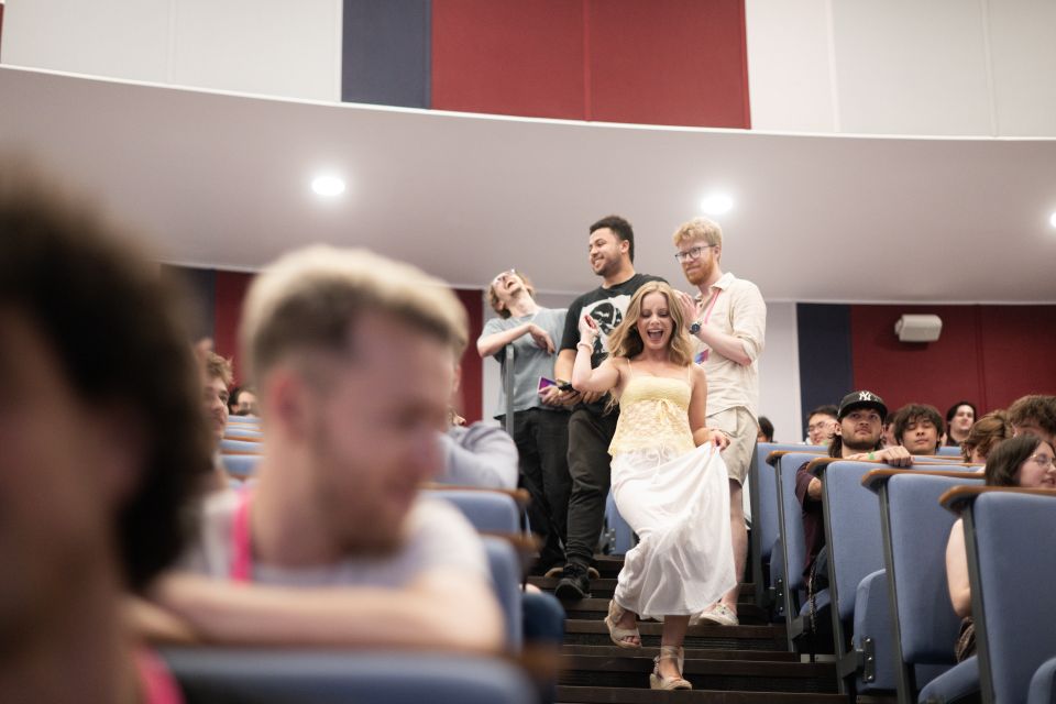 A young woman in a white dress energetically descends lecture theatre stairs, surrounded by smiling people seated.