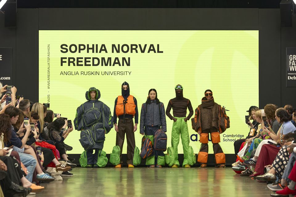 Five models showcase colorful, futuristic outfits on a runway, with an audience and a large screen in the background displaying text.