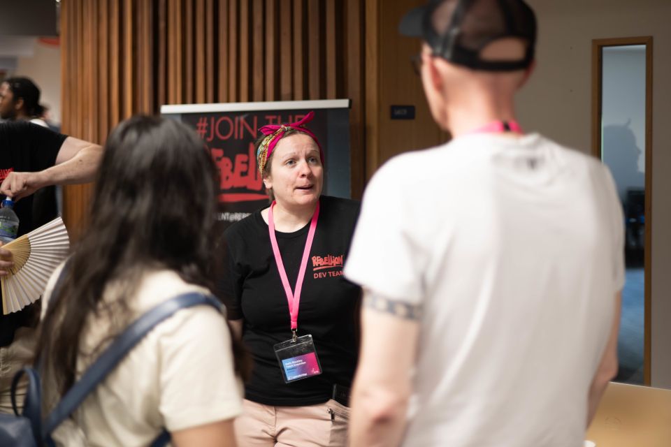 A Rebellion rep with a pink lanyard talks to two people at a booth with a "#JoinTheRebellion" sign in the background.