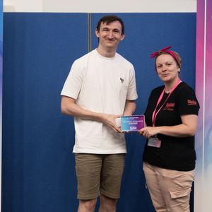 Two people stand smiling, holding an award. They are between two banners with various sponsor logos at a student games festival.