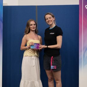 Two women smiling and holding an award at a gaming festival, standing between two sponsor banners.