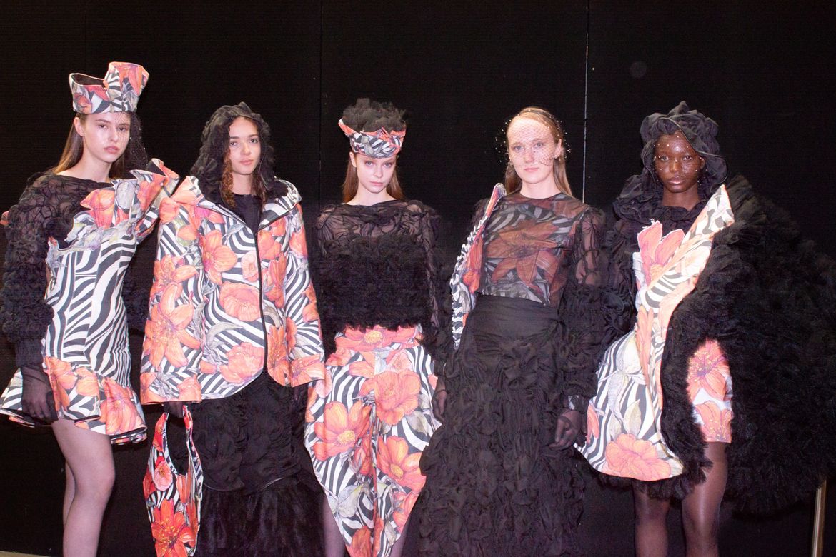 Five models in bold floral and zebra-patterned outfits with dramatic headpieces pose confidently against a dark background.