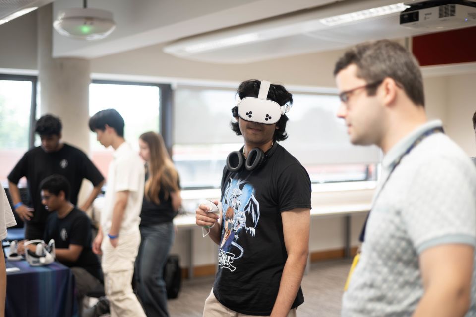 A young man wearing a VR headset and headphones stands in a room with others, holding a controller.