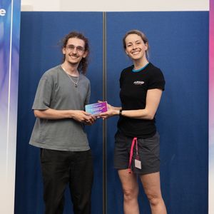 Two people smiling and holding an award between them at a student games festival, standing in front of banners with various sponsor logos.