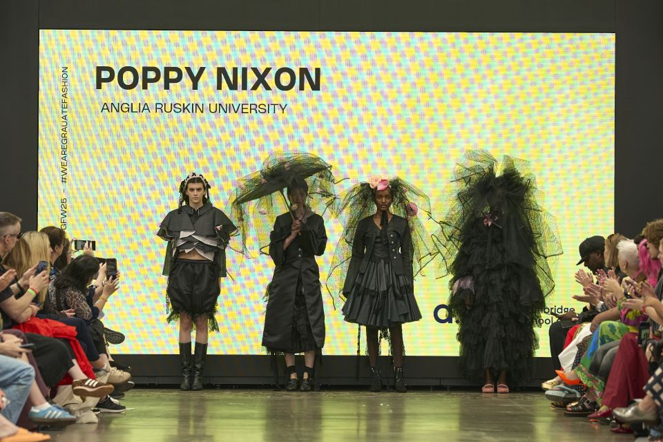 Four models in avant-garde black outfits stand on a runway, with a bright yellow backdrop displaying "Poppy Nixon, Anglia Ruskin University."