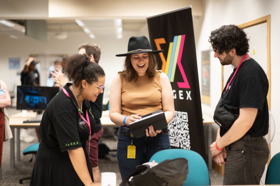 Three people engaging in conversation at an event, with a colourful banner in the background. One is wearing a hat and holding a tablet.
