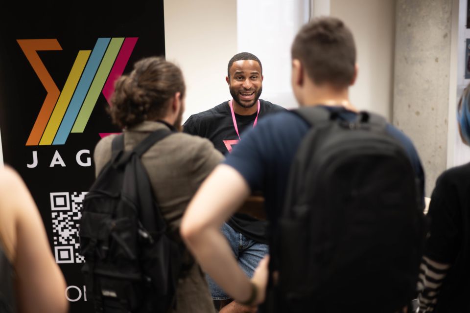 A man speaks enthusiastically to a group of people at an event, standing in front of a Jagex banner with colourful graphics and a QR code.